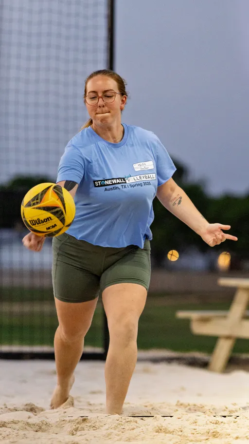 A volleyball player in a light blue team shirt and green shorts is serving a yellow and black volleyball on a sandy outdoor court, with a net and picnic table visible in the background.