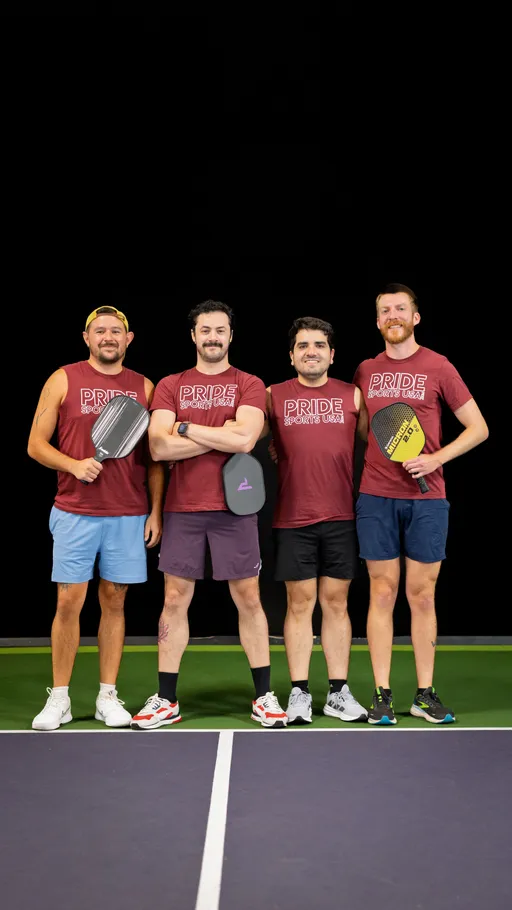 Four pickleball players stand side by side on an indoor pickleball court, each holding a paddle and wearing matching maroon "Pride Sports USA" shirts, athletic shorts, and sneakers.