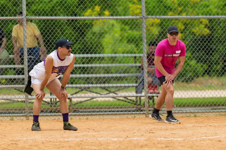 Two kickball players are positioned on a sandy field, both crouching and ready for action; one wears a white jersey with a colorful design and purple accessories, while the other wears a bright pink shirt and dark shorts. A chain-link fence and benches with spectators are visible in the background.