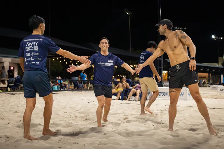 On a lit sand volleyball court at night, several players exchange high-fives between points, smiling as they move across the sand. Most wear matching navy team shirts and shorts with sponsor logos, while one player wears black shorts, a cap, and a watch; spectators and other competitors relax on benches under string lights in the background.
