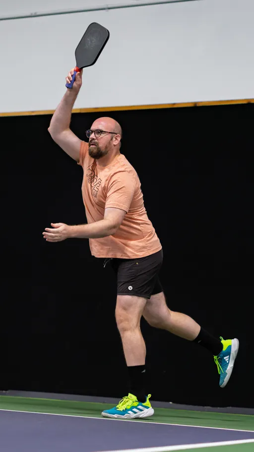 A pickleball player wearing a peach t-shirt, black shorts, black socks, and blue athletic shoes is in mid-action, swinging a paddle overhead on an indoor court. The background is a dark wall, emphasizing the motion and focus of the game.