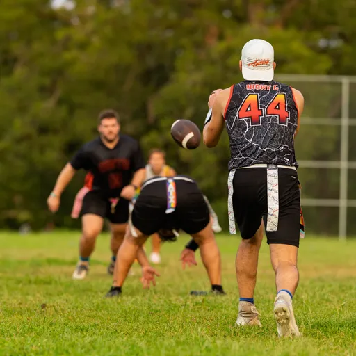 On a grassy park field, ATX Heat (Orange) players run a flag football drill; a player in a black jersey with number 44 and a cap winds up to pass while teammates sprint routes and defenders close in. The group wears cleats and flag belts, with a chain‑link backstop and trees in the background, capturing practice for Gay Bowl 2025.