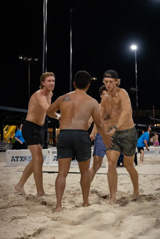 On a lit outdoor sand volleyball court at night, four players huddle with hands in for a team cheer between points. They wear athletic shorts—one in camo shorts with a backwards cap—and other matches are visible on nearby courts.