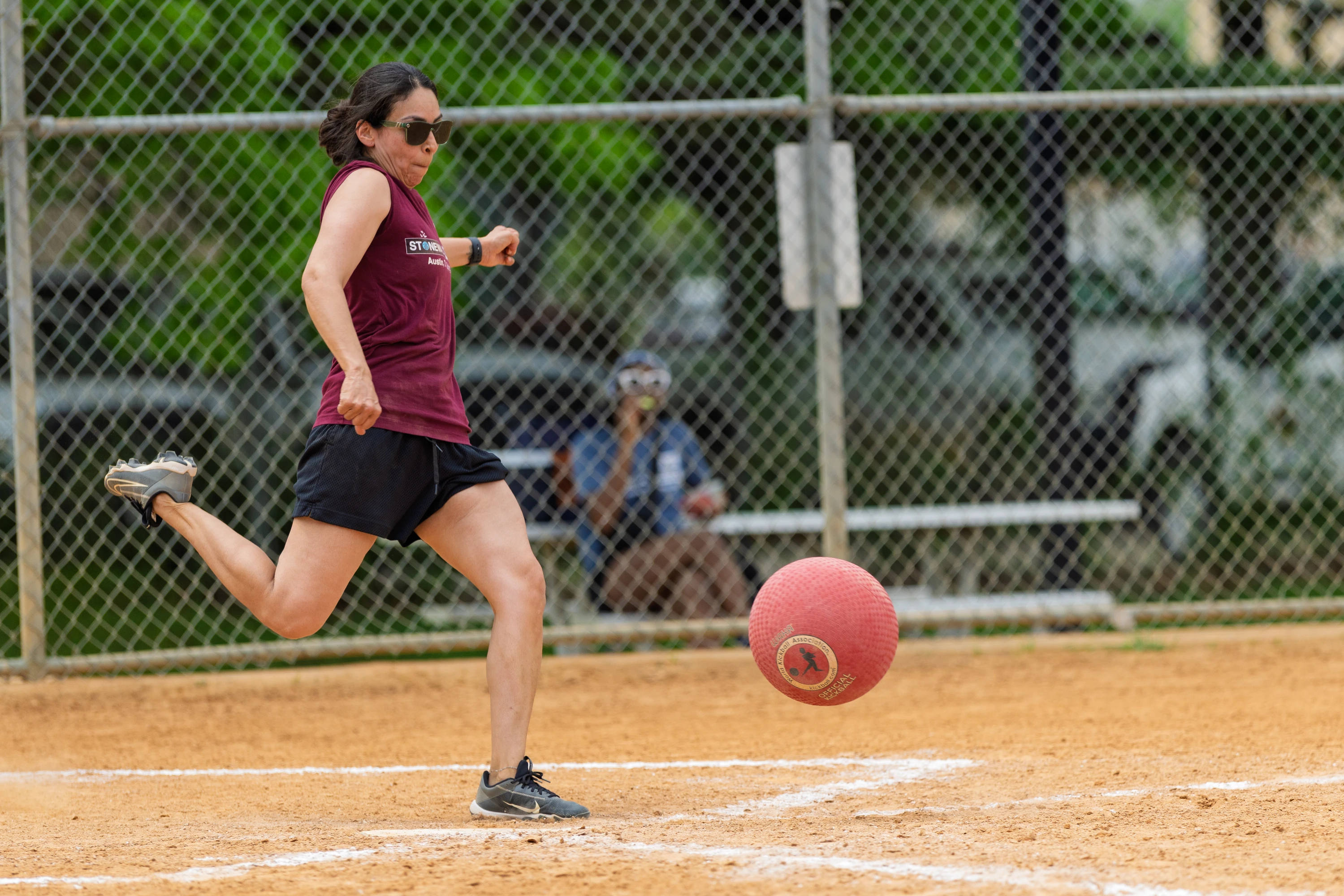 A player wearing a maroon sleeveless shirt, black shorts, and sunglasses is kicking a red kickball on a dirt field, with a chain-link fence and a seated person visible in the background.