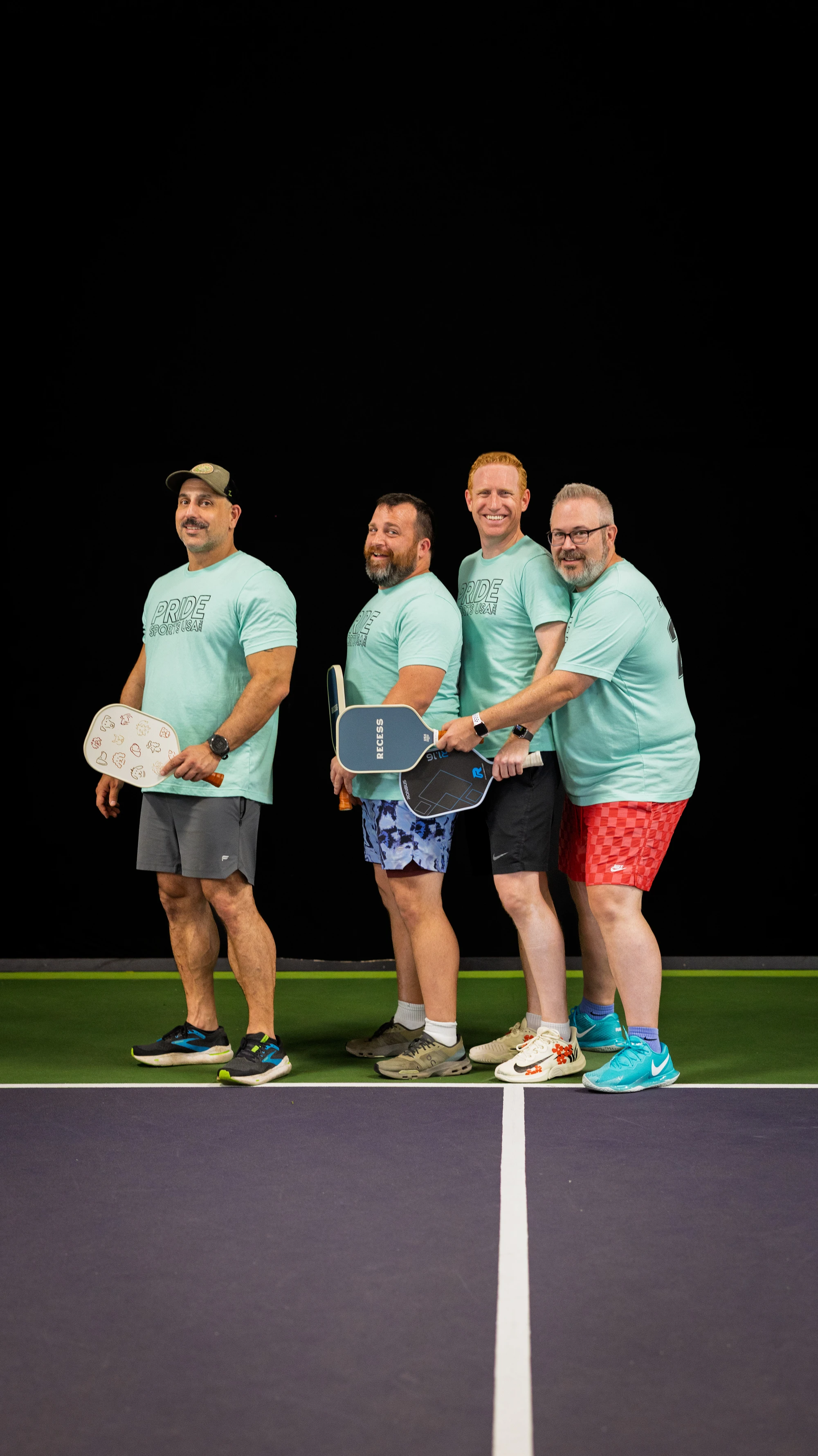 Four pickleball players stand side by side on an indoor court, each holding a paddle and wearing light blue matching "Pride Pickleball" t-shirts, athletic shorts, and sneakers. They are posing and smiling, ready for the game.