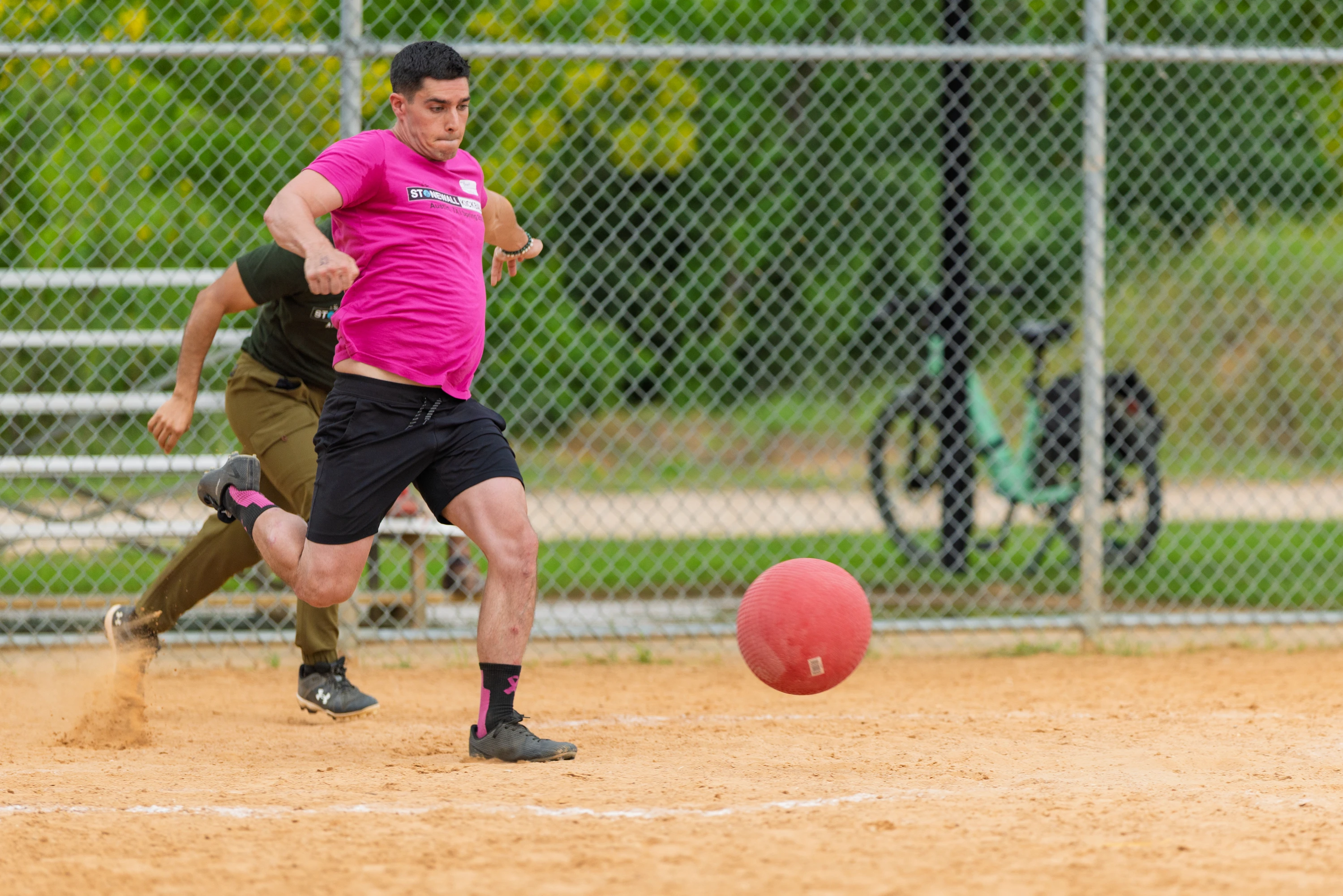 A player in a bright pink shirt and black shorts is kicking a red kickball on a dirt field, while another player in an olive green shirt runs behind them; a chain-link fence and a bicycle are visible in the background.