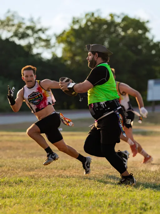 On a sunlit grassy field, ATX Heat (Black) players practice for Gay Bowl 2025: a ball-carrier in a neon green pinnie, black leggings, cap, and flag belt runs with the football while defenders in ATX Heat crop jerseys, cleats, and flag belts sprint in pursuit, reaching to pull a flag amid trees in the background.