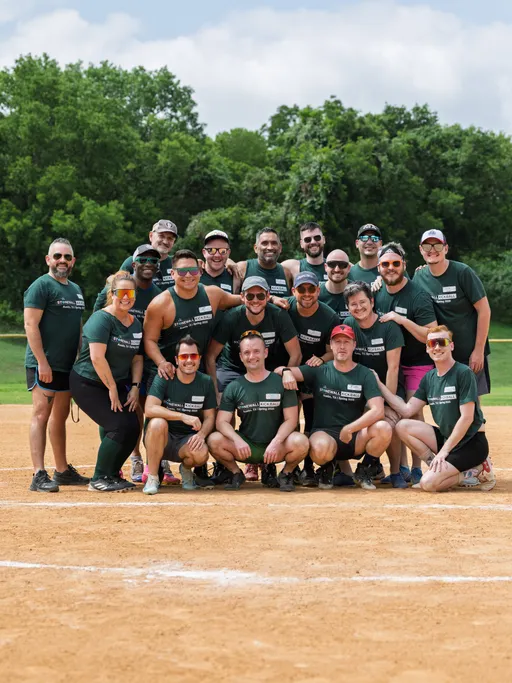 A group of kickball players pose together on a kickball field, all wearing matching dark green team shirts and various athletic gear, with trees and grass visible in the background. The team appears to be celebrating or commemorating a game during the Stonewall Kickball Spring 2025 season.