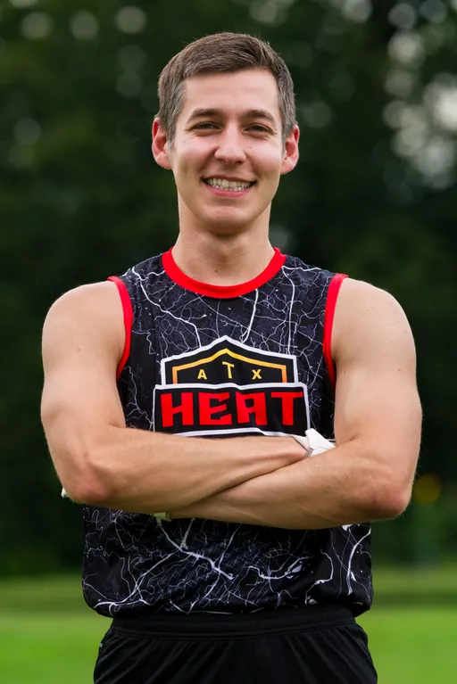 An ATX Heat (Orange) player poses on a grassy practice field with arms crossed, wearing a black sleeveless jersey with red trim and a marble pattern that reads “ATX HEAT,” with blurred trees in the background during Gay Bowl 2025 prep.