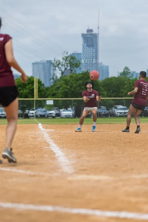Several kickball players wearing matching maroon shirts and athletic shorts are on a dirt field, with one player preparing to catch a red kickball while others move into position; a city skyline and parked cars are visible in the background.