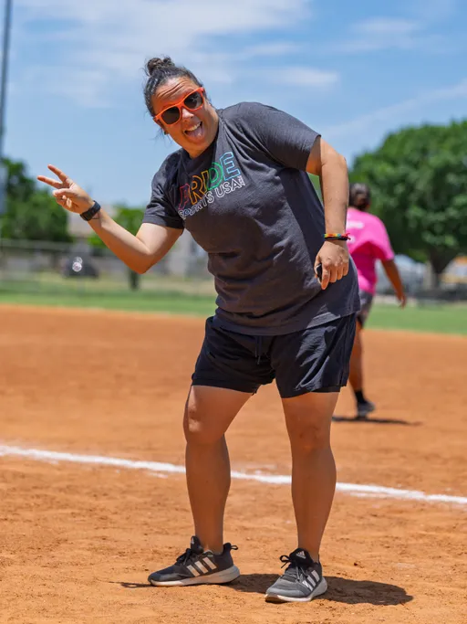 A player on the kickball field poses near a baseline, wearing a "Pride Sports USA" t-shirt, black shorts, athletic shoes, orange sunglasses, and a rainbow wristband, making a peace sign with one hand. Another player in a pink shirt can be seen in the background.