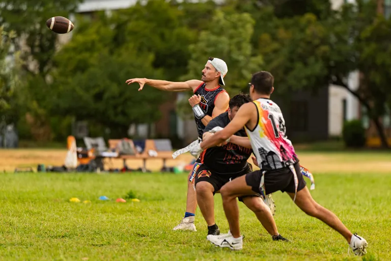 On a grassy field, an ATX Heat (Orange) quarterback in a black-and-orange sleeveless jersey and backward cap throws a football as two defenders in multicolored flag-football jerseys close in, reaching for the flag belt. Cones, benches, and trees frame the practice area during a Gay Bowl 2025 prep scrimmage.