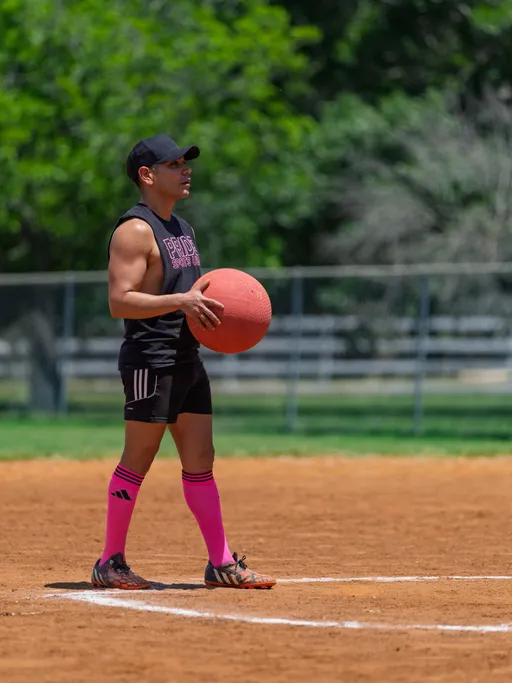 A player stands on a kickball field holding a large red kickball, wearing a black sleeveless "Pride Kickball" shirt, black shorts, and knee-high pink socks, preparing to pitch.