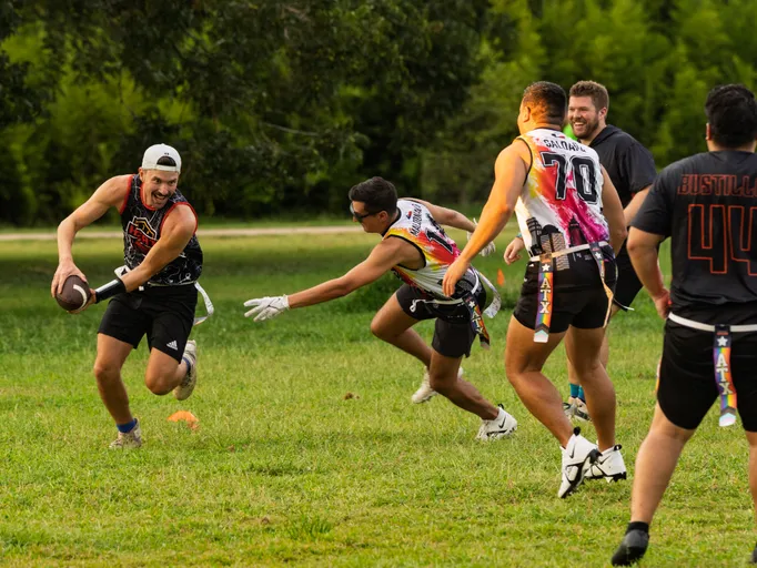 On a grassy park field, ATX Heat (Orange) flag football players scrimmage for Gay Bowl 2025 practice: a ball carrier in a black sleeveless jersey, cap, and gloves sprints left with the football while defenders in colorful ATX jerseys and flag belts reach to pull the runner’s flag near field cones and trees.