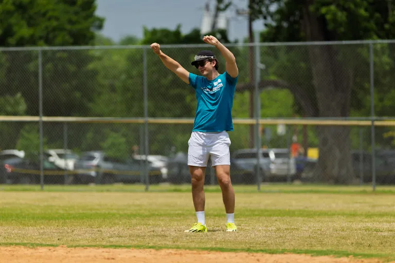 A player in a teal shirt, white shorts, bright shoes, and a visor is standing on the kickball field with both arms raised, possibly celebrating or signaling during a game. The scene takes place outdoors with a chain-link fence and trees in the background.