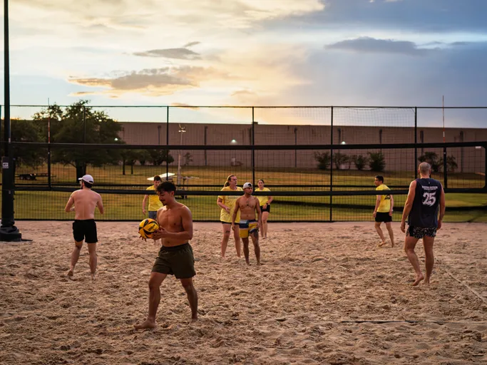 Several volleyball players are on a sand court at sunset, some wearing yellow team shirts while others wear shorts and tank tops or athletic tops; one player in the foreground is holding a volleyball as the game resumes.