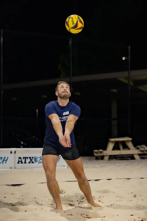A sand volleyball player in a navy T-shirt with a white logo and black shorts braces for a forearm pass, arms together as a yellow‑and‑purple ball descends; the action takes place on a lit night sand court with fencing, signage, and a picnic table in the background.