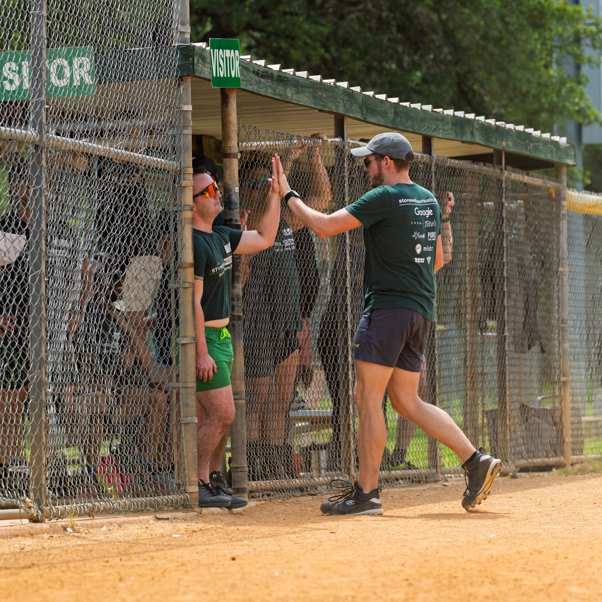A player wearing a green shirt and shorts is giving a high five to another player standing in the dugout area next to the kickball field, with both individuals appearing cheerful and engaged in the game. The environment includes fencing, signage that reads "VISITOR", and other players and trees in the background.