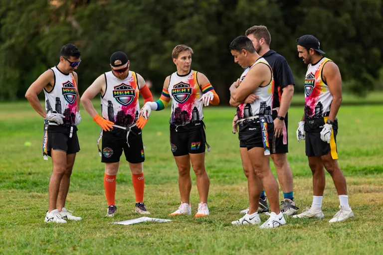 On a grassy practice field, ATX Heat (Orange) players huddle around a play sheet on the ground, discussing strategy during Gay Bowl 2025 prep. They wear matching white jerseys with the HEAT logo and orange/pink graphics, black shorts with flag belts, cleats, and assorted accessories like gloves, a cap, and an armband, with trees lining the background.