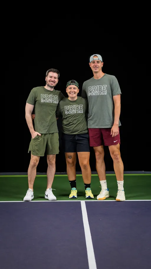 Three pickleball players stand together on an indoor court, all wearing "Pride Sports USA" shirts and athletic shorts, with two wearing caps and one wearing a headband.