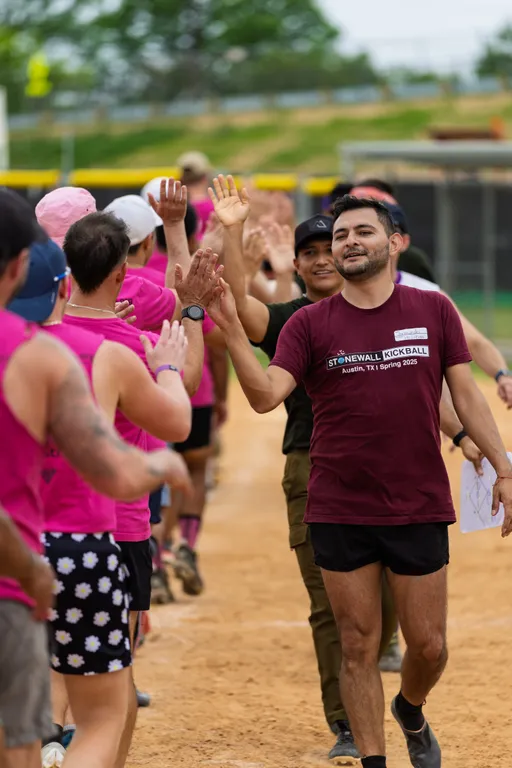 Players are lined up on a kickball field, exchanging high fives; most are wearing athletic gear, with one player in the foreground wearing a maroon "Stonewall Kickball Austin, TX Spring 2025" shirt and black shorts, while others sport bright pink shirts. The scene takes place on a dirt field with green grass and a yellow fence visible in the background.