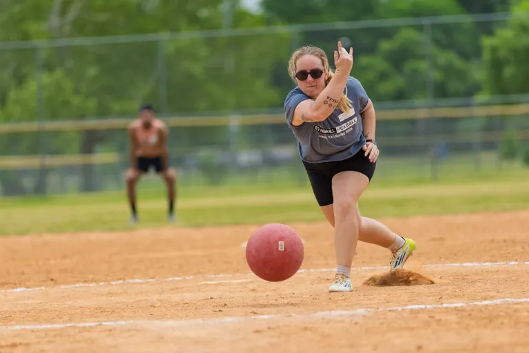 A player wearing a gray Stonewall Kickball t-shirt, black shorts, and sunglasses is in motion, pitching a red kickball on a dirt field, with another player crouched in the outfield ready to react. The scene takes place outdoors, surrounded by green trees and athletic field fencing.