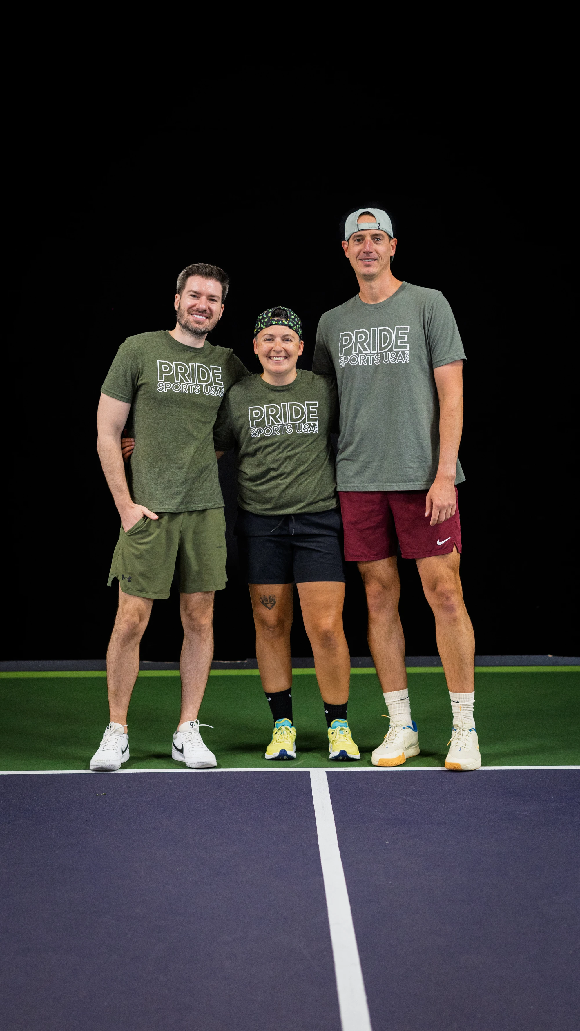 Three pickleball players stand together on an indoor court, all wearing "Pride Sports USA" shirts and athletic shorts, with two wearing caps and one wearing a headband.
