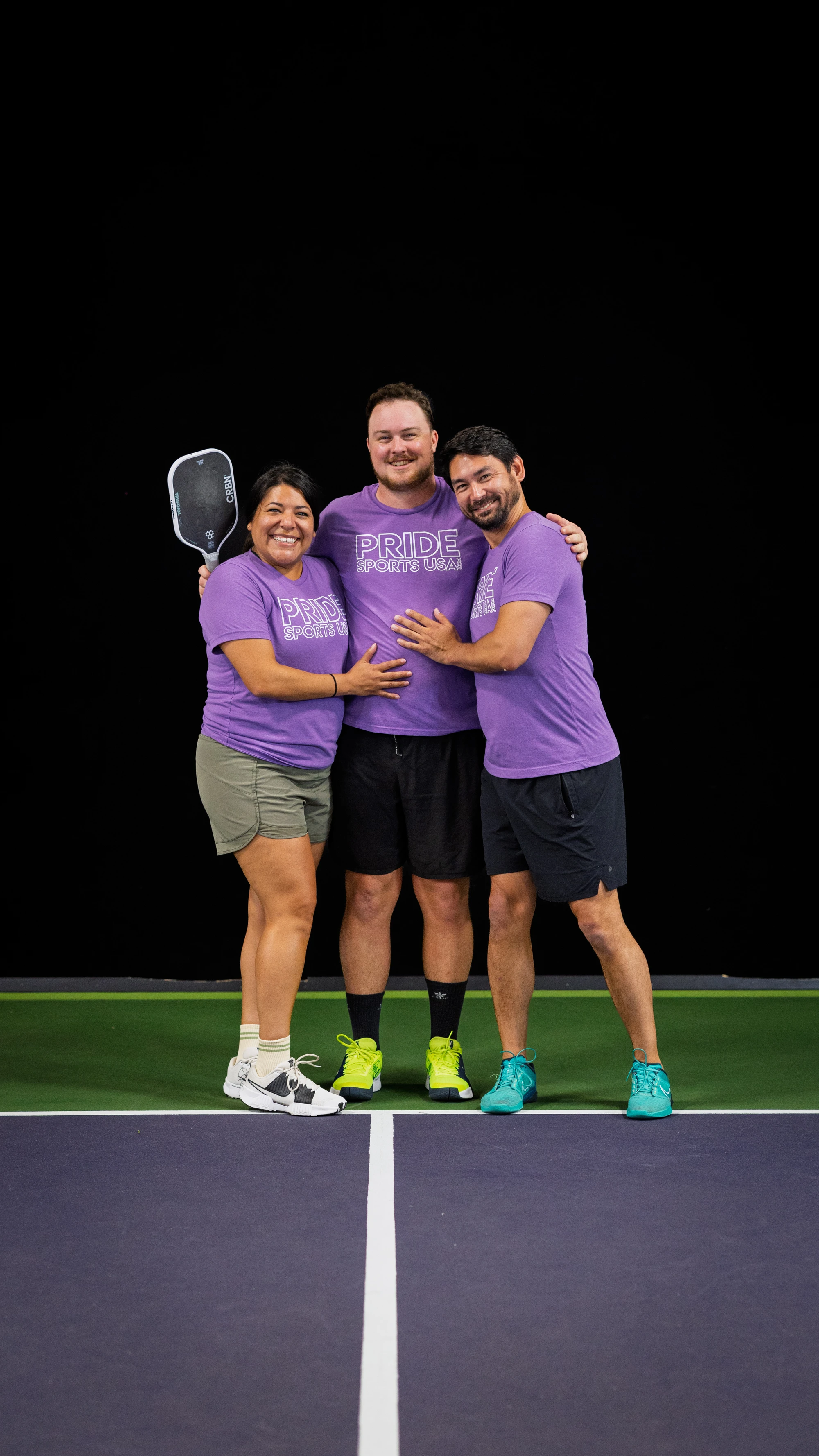 Three pickleball players stand together smiling on an indoor court, each wearing matching purple "Pride Sports USA" shirts, with one person holding a pickleball paddle.