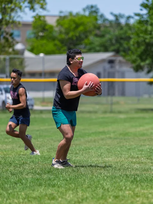 A player on a grassy kickball field prepares to throw a red kickball, while another player runs in the background; both wear dark shirts and athletic shorts.