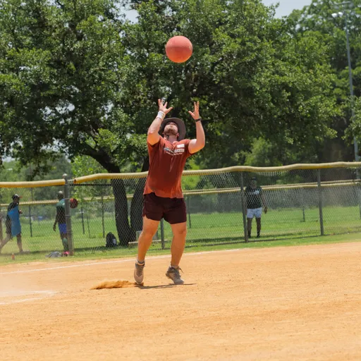A kickball player wearing a wide-brimmed hat, maroon shirt, and shorts prepares to catch a kickball descending from above on a sunny kickball field, with a fence and trees visible in the background.