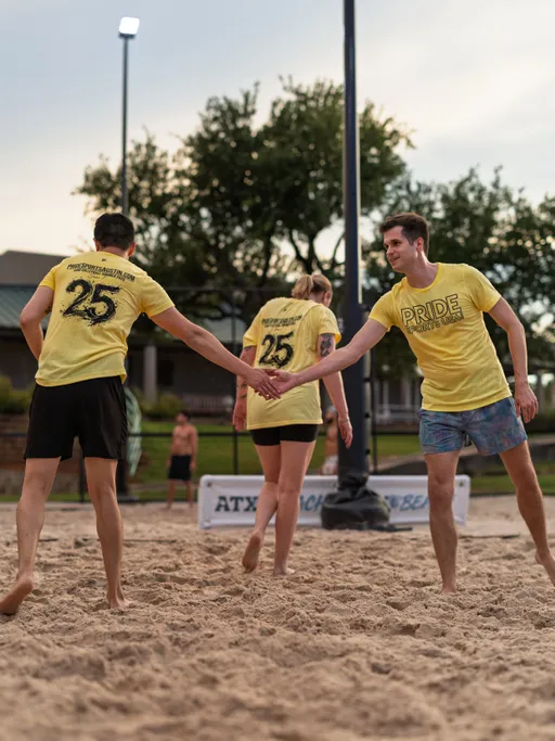 Three volleyball players wearing matching yellow "Pride Sand Volleyball" shirts are on a sand court, with two of them giving each other a high five and another walking nearby; the scene takes place outdoors with sports equipment and a fence visible in the background.
