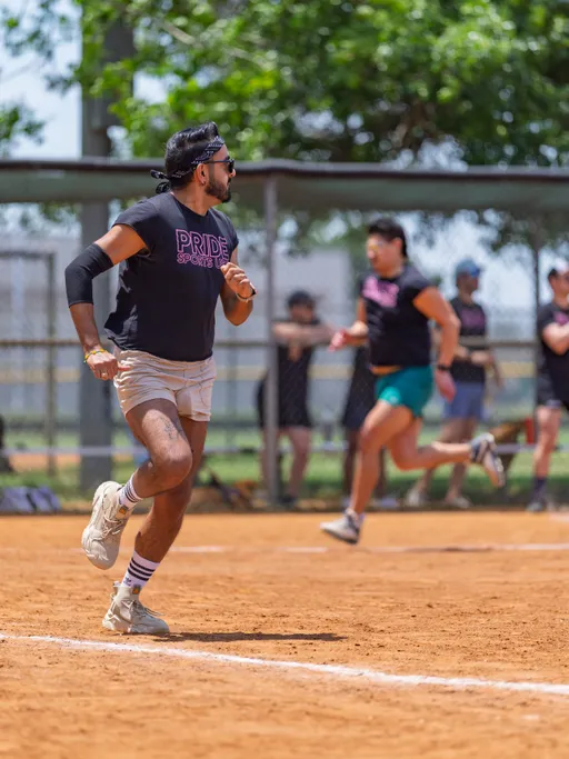 Several players are running on a dirt kickball field, all wearing black PRIDE SPORTS t-shirts and athletic shorts, with one player in the foreground focused on the game and wearing a headband and arm sleeve. The background shows more participants and a fence, with trees providing some shade.