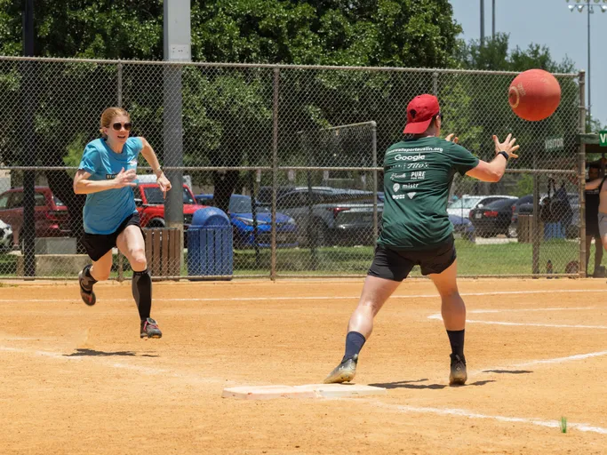 A player wearing a blue shirt and black shorts is running toward a base while another player in a green shirt and red cap prepares to catch a red kickball near the base on a dirt kickball field, with a chain-link fence and parked cars in the background.
