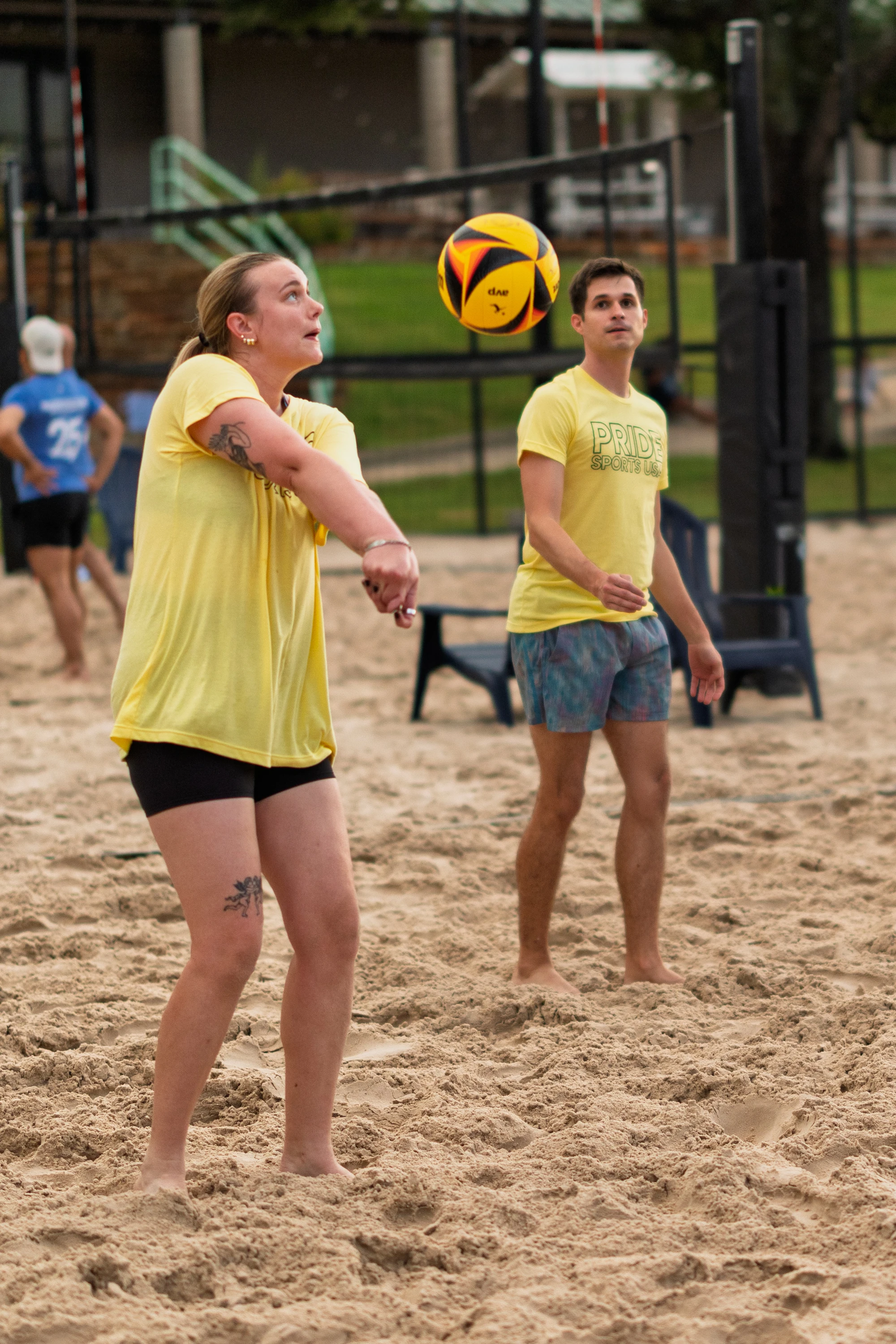 Two volleyball players in yellow shirts, one with "PRIDE" printed on it, are on a sandy outdoor court; one player is preparing to bump a yellow and black volleyball while the other stands nearby watching the play.