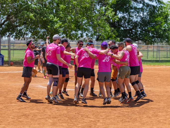 A group of kickball players wearing matching bright pink shirts with numbers on the back are gathered in a huddle on a dirt kickball field, some jumping and cheering while surrounded by trees and a chain-link fence.