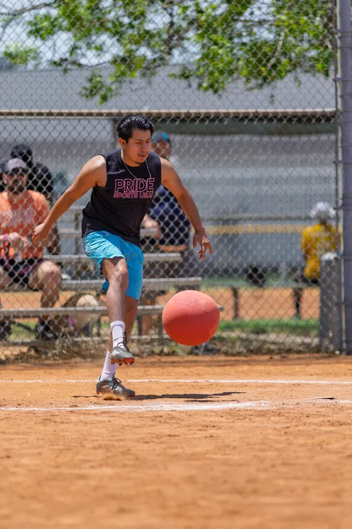 A player wearing a "Pride Sports USA" sleeveless shirt and blue shorts is preparing to kick a red kickball on a dirt field, with spectators seated on bleachers in the background and a chain-link fence behind them.