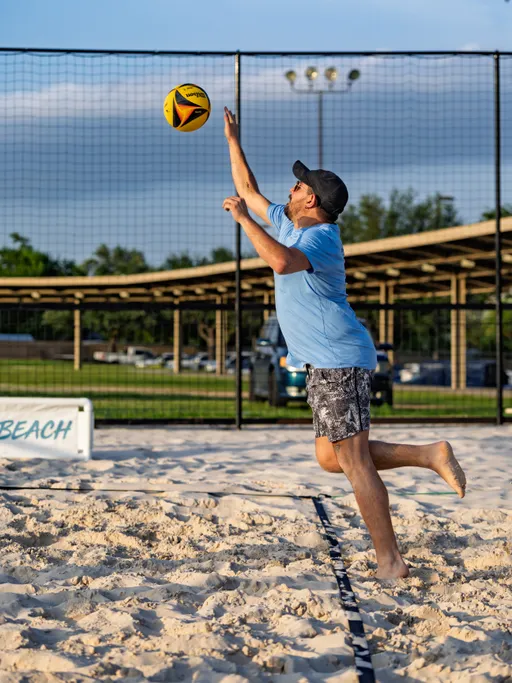 A volleyball player wearing a light blue shirt, patterned shorts, and a dark cap is jumping barefoot to serve a volleyball on a sandy outdoor court, with a grassy area and parked cars visible in the background.