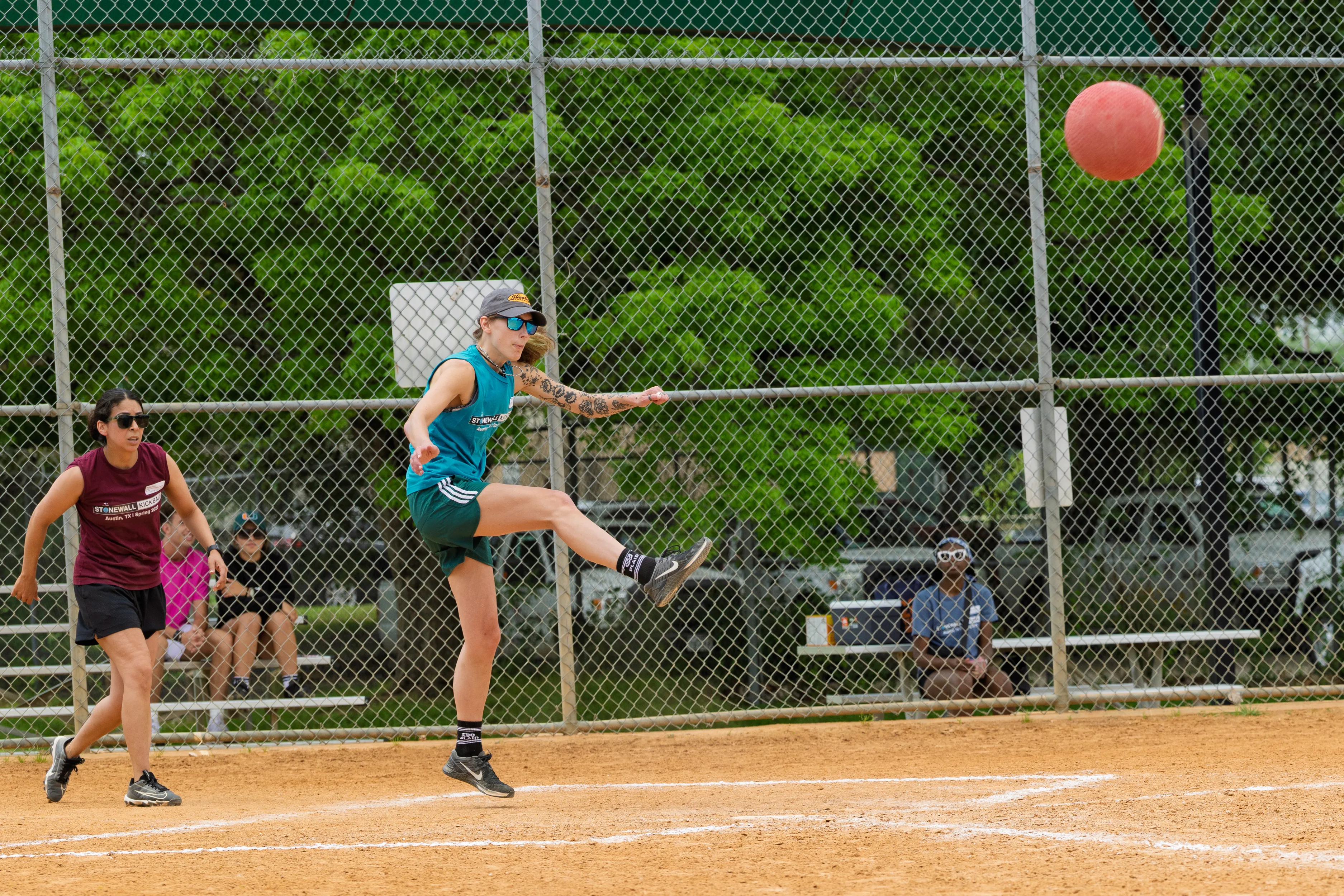 A player in a teal jersey, green shorts, cap, sunglasses, and knee-high socks launches a red kickball with a high kick near home plate on a dusty kickball field. A maroon-clad defender hustles in as a few spectators watch from bleachers behind a chain-link fence and leafy trees.