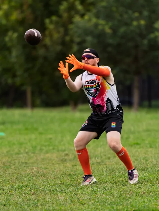 On a grassy park field, an ATX Heat (Orange) player reaches to catch a football during Gay Bowl practice, wearing a white “HEAT” team tank with cityscape graphic, black shorts, bright orange gloves and knee-high socks, a black cap, and sunglasses, with trees blurred in the background.
