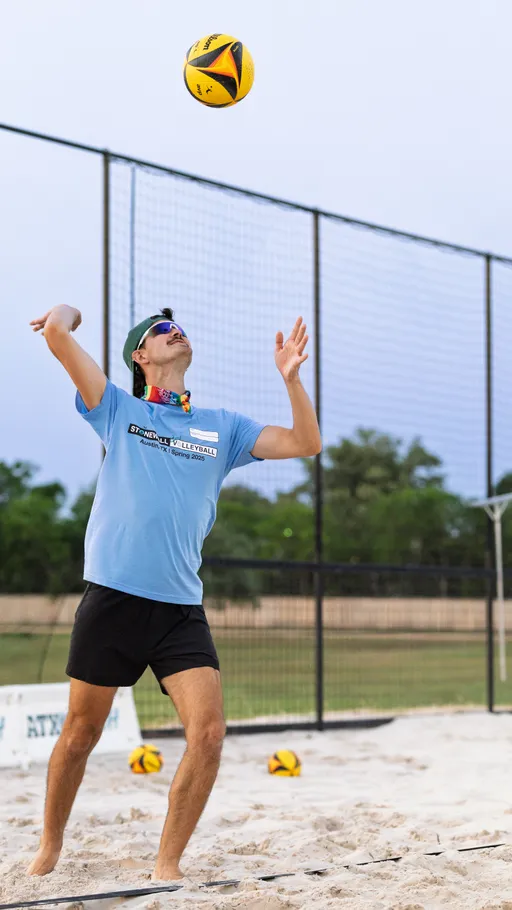 A volleyball player on a sand court prepares to serve, tossing a volleyball overhead. The player is wearing a light blue "Stonewall Volleyball" t-shirt, black shorts, sunglasses, a cap, and a colorful bandana, with additional volleyballs and a net visible in the background.