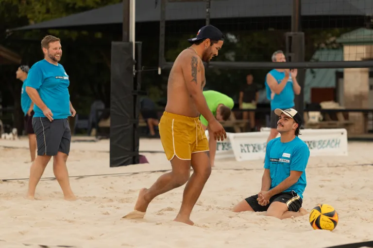 On an outdoor sand volleyball court, a player in yellow shorts and a backwards cap walks past a teammate in a blue team shirt and cap who is kneeling in the sand beside a yellow-and-black volleyball, with other blue-shirted players and the net visible in the background.
