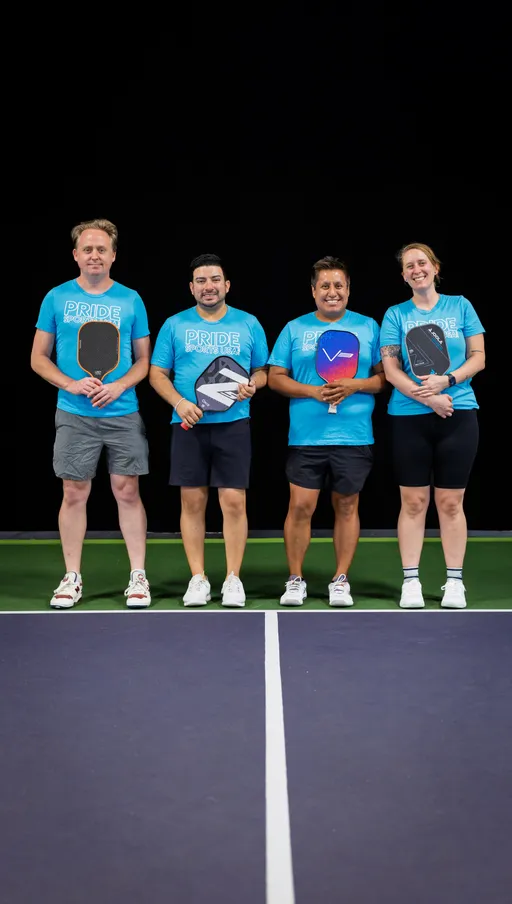 Four pickleball players stand on an indoor court, each holding a pickleball paddle and wearing matching blue "Pride Pickleball" shirts, shorts, and athletic shoes. They are posed side by side and smiling, ready for the game.