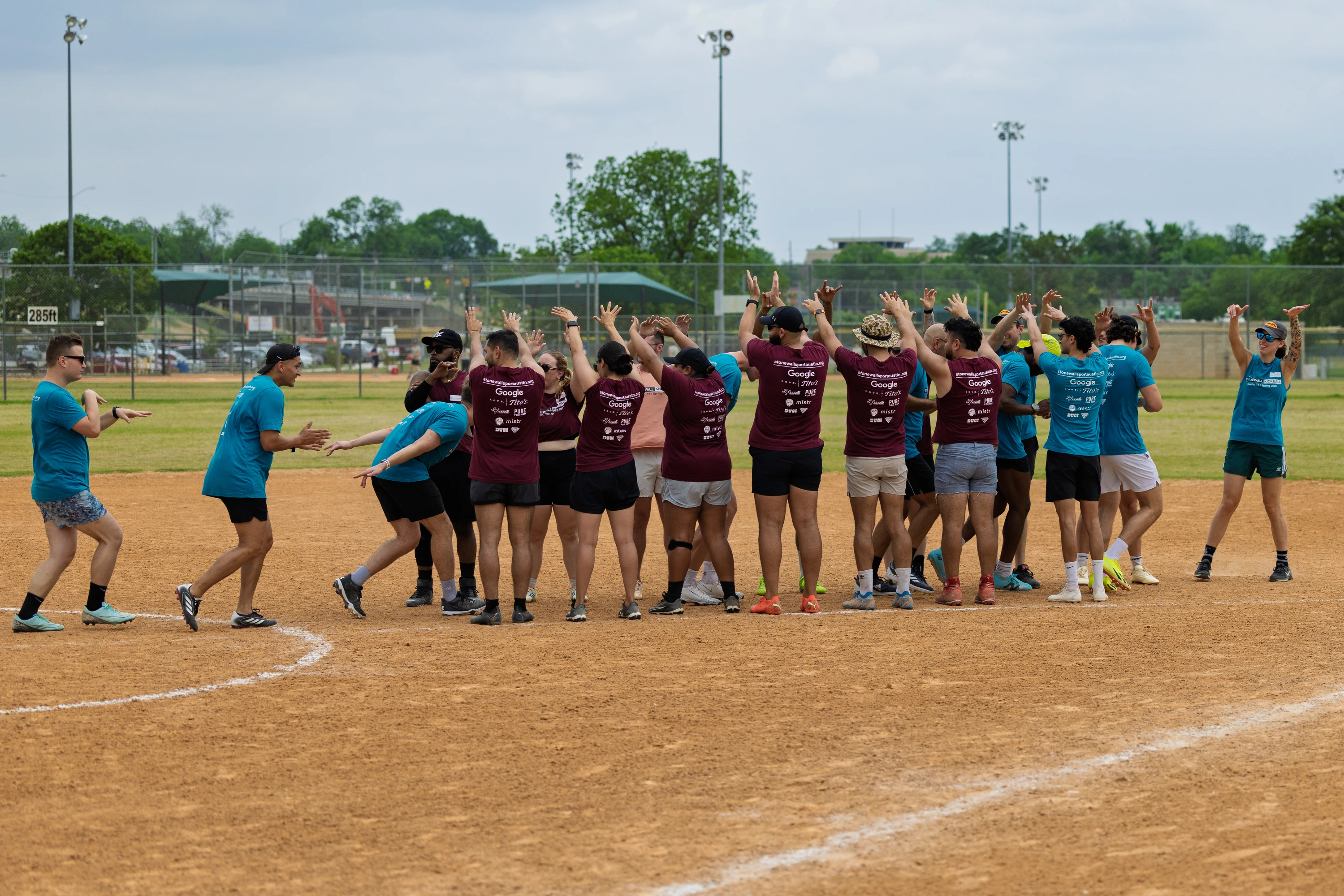 A group of kickball players stands in a circle on a dirt field, many with arms raised or clapping, wearing matching maroon or teal team shirts and athletic shoes, while engaging in a group cheer or huddle before a game.