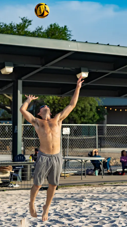 A volleyball player in gray shorts jumps on a sand court with an arm extended upward to serve or hit a yellow and black volleyball, with spectators and a shaded seating area in the background.