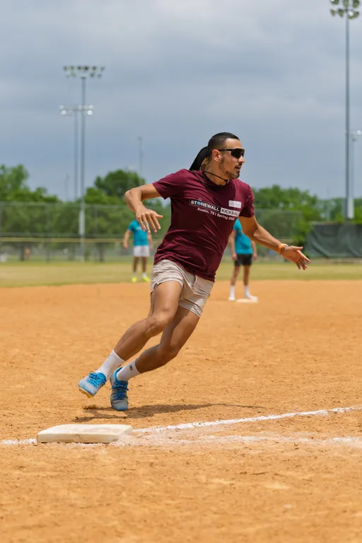 A player wearing a maroon "Stonewall Kickball" shirt, light shorts, blue shoes, and sunglasses is running past a base on a sandy kickball field, with other players and field lights visible in the background.
