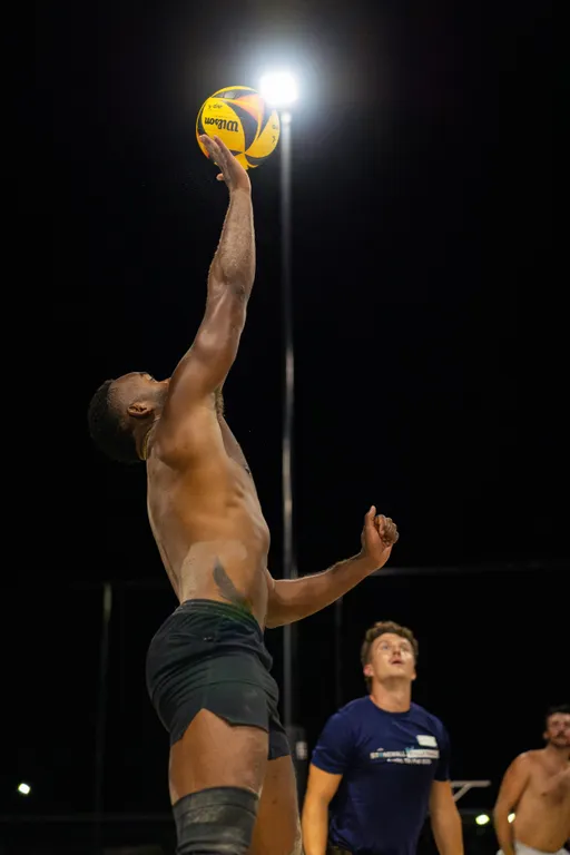 On an outdoor sand volleyball court at night, a player in dark shorts jumps and stretches one arm to make a one-handed play on a yellow-and-blue volleyball under bright lights. Other players on the court watch the action from the sand.