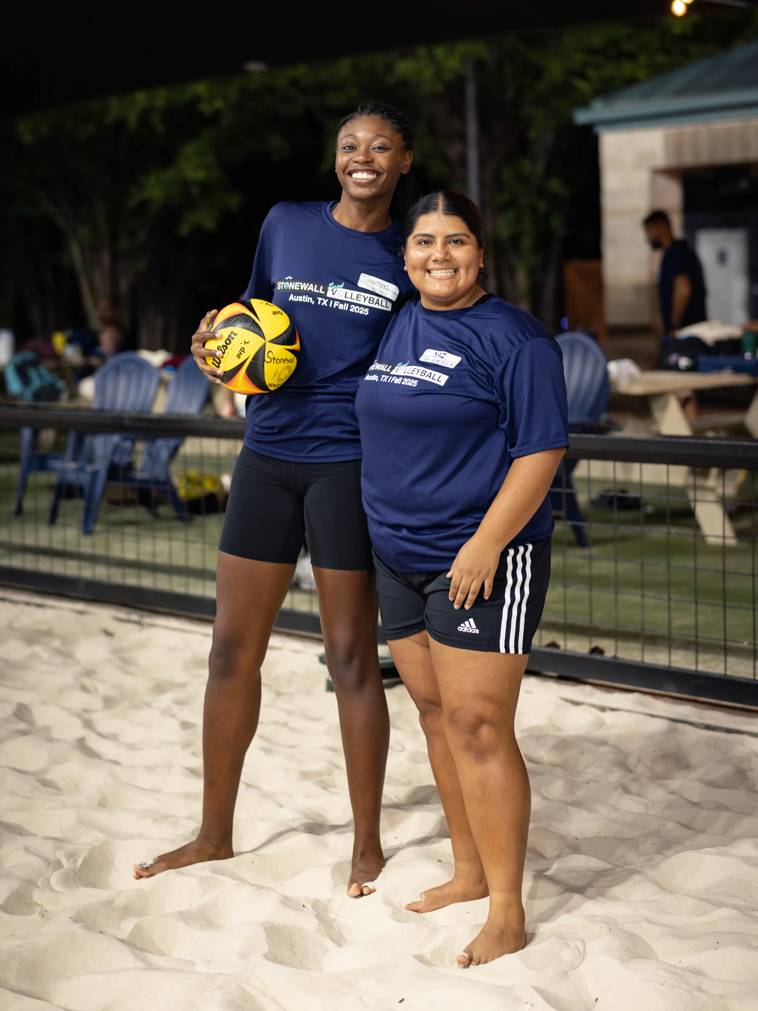 Two players stand barefoot on a lit sand volleyball court, smiling side by side. They wear navy Stonewall Volleyball shirts and black shorts; one holds a yellow-and-black volleyball, with fencing, benches, and lounge chairs in the background.