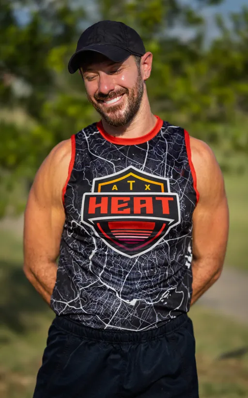 An ATX Heat player stands on a sunny practice field, smiling with hands behind their back, wearing a black cap and a black sleeveless team jersey with red trim and a bold “ATX HEAT” logo over a map-style pattern; blurred trees frame the outdoor summer scene as the team prepares for Gay Bowl 2025.