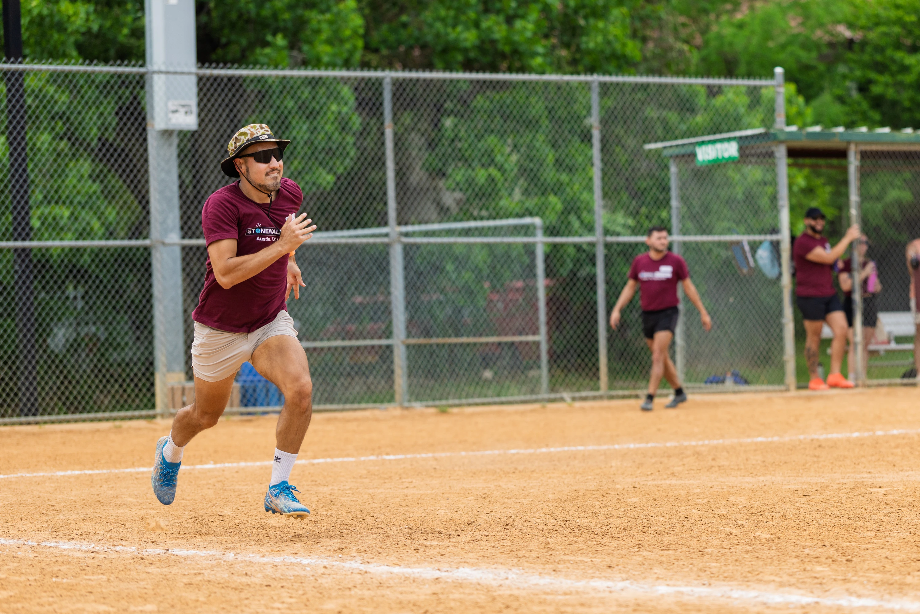 A kickball player wearing a maroon Stonewall team shirt, light shorts, blue cleats, sunglasses, and a bucket hat is running on the dirt field, with other team members and a chain-link fence visible in the background.