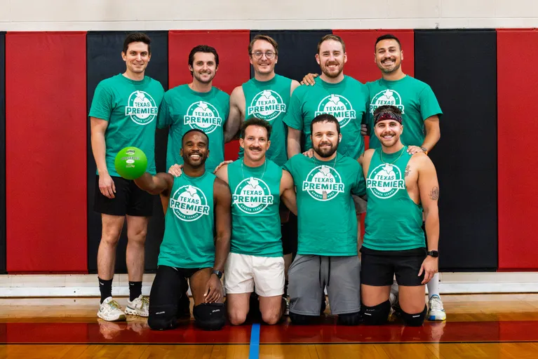 A group of TPSL Dodgeball players pose for a team photo in a gym, standing and kneeling in two rows against red-and-black wall padding. They wear matching green “Texas Premier” shirts with shorts and athletic shoes; some have knee pads or headbands, and one player holds a green dodgeball.